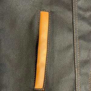 Puede incluir: Primer plano de una chaqueta vaquera azul oscuro con un detalle de bolsillo vertical rectangular de cuero marr&oacute;n claro. El bolsillo est&aacute; delineado con costuras marrones claras, y la chaqueta tiene costuras marrones claras adicionales a lo largo de las costuras.