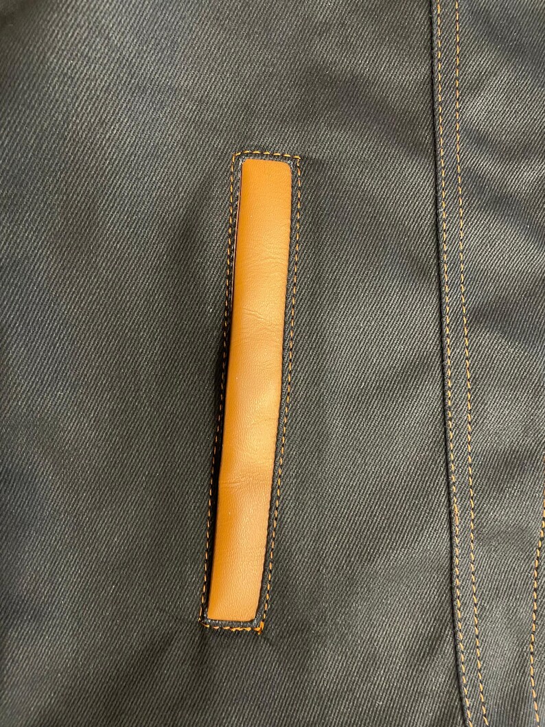 Puede incluir: Primer plano de una chaqueta vaquera azul oscuro con un detalle de bolsillo vertical rectangular de cuero marr&oacute;n claro. El bolsillo est&aacute; delineado con costuras marrones claras, y la chaqueta tiene costuras marrones claras adicionales a lo largo de las costuras.