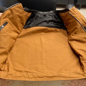 K&ouml;nnte beinhalten: Eine braune Leder-Motorradjacke mit schwarzem Futter. Die Jacke ist ge&ouml;ffnet und flach gelegt, so dass das Innere der Jacke sichtbar ist.