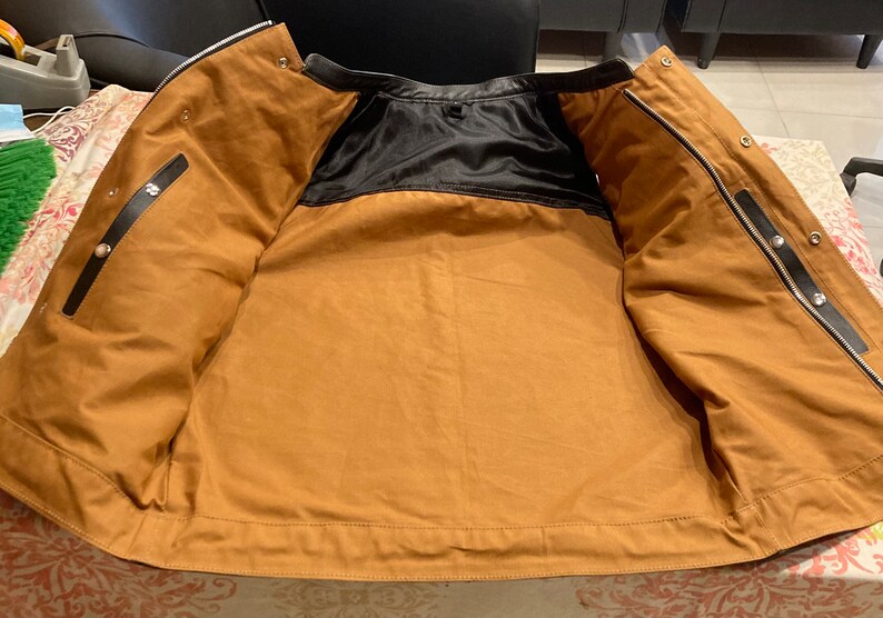 K&ouml;nnte beinhalten: Eine braune Leder-Motorradjacke mit schwarzem Futter. Die Jacke ist ge&ouml;ffnet und flach gelegt, so dass das Innere der Jacke sichtbar ist.
