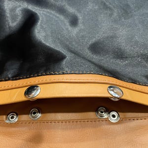 Puede incluir: Primer plano del interior de un bolso de cuero. El bolso presenta un exterior de cuero marr&oacute;n con un forro de sat&eacute;n negro. Los cierres de presi&oacute;n plateados aseguran la apertura del bolso. Las costuras son visibles en los bordes.