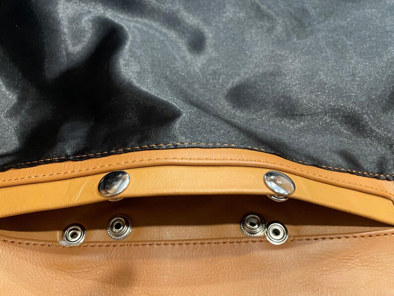 Puede incluir: Primer plano del interior de un bolso de cuero. El bolso presenta un exterior de cuero marr&oacute;n con un forro de sat&eacute;n negro. Los cierres de presi&oacute;n plateados aseguran la apertura del bolso. Las costuras son visibles en los bordes.