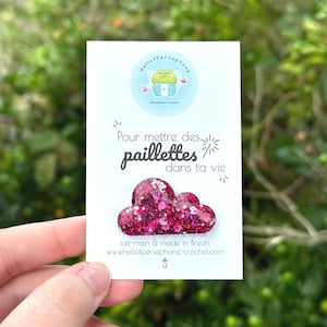 May include: A handmade cloud-shaped brooch with a glittery pink and magenta design. The brooch is on a white card with the text "Pour mettre des paillettes dans ta vie" and the website "www.hellofpersephone-crochet.com".