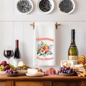 Op de afbeelding: Een witte keukendoek met de woorden "Manman Poto Mitan" in rode letters en een bloemenontwerp. De handdoek hangt aan een houten stang boven een tafel met wijn, kaas en druiven.