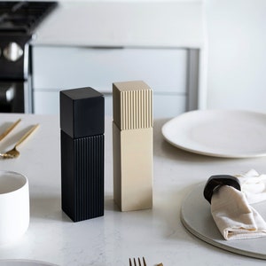 Peut inclure: Deux salières et poivrières sur un comptoir blanc. Les salières sont en céramique et ont un design côtelé. La salière de gauche est noire et la salière de droite est beige.