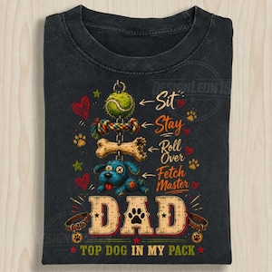 Könnte beinhalten: Dunkelgraues T-Shirt mit einem Hunde-Design. Das Shirt zeigt ein Hundespielzeug, einen Knochen und einen blauen Cartoon-Hund. Der Text enthält "Sit, Stay, Roll Over, Fetch Master" und "DAD, Top Dog in My Pack."