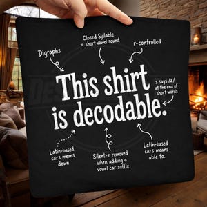 Puede incluir: Un cuadrado de tela negro con el texto blanco This shirt is decodable. Flechas señalan notas lingüísticas que incluyen Digraphs, Closed Syllable, r-controlled, s says /z/, Latin-based cars means down, Silent-e removed y Latin-based cars means able to.