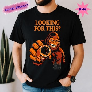 Puede incluir: Camiseta negra con el texto "LOOKING FOR THIS?" y una ilustración de dibujos animados de un Bigfoot sosteniendo una llave de vaso de 10 mm. El Bigfoot es marrón y naranja, con una expresión sonriente.