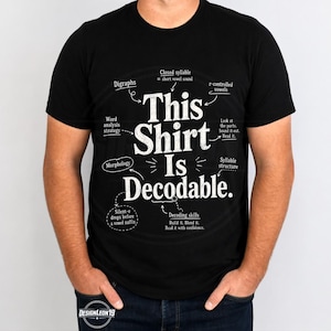 Op de afbeelding: Een zwart t-shirt met korte mouwen en witte tekst die This Shirt Is Decodable leest. Het ontwerp bevat verschillende educatieve taalkundige termen en pijlen die naar de centrale tekst wijzen, zoals Digraphs, Morphology en Decoding skills.