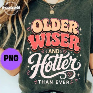 May include: Dark green t-shirt with the text "OLDER WISER AND Hotter THAN EVER" in a retro font. The design includes hearts and stars. The shirt is a relaxed fit.
