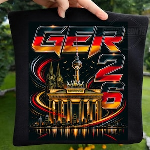 May include: A black fabric item featuring a graphic print of the Brandenburg Gate, the Berlin TV Tower, and Cologne Cathedral. The design includes the text GER 26 in bold lettering filled with the black, red, and gold colours of the German flag.