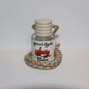 Pode incluir: Uma pequena garrafa de vidro com um cordão bege pendurado no topo. A garrafa tem uma tampa de madeira branca e um rótulo que diz "Spiced Apple Cider" com uma imagem de um copo de cidra, uma maçã e paus de canela.