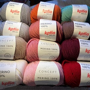 May include: A collection of 12 balls of yarn in various colours, including pink, red, brown, beige, and green. Each ball has a label that reads "Merino 100% Best Basics Katia 100% You".