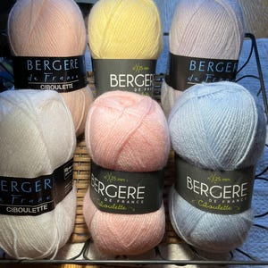 Può includere: Sei gomitoli di lana in vari colori pastello, tra cui rosa, giallo, azzurro e bianco. Ogni gomitolo ha un'etichetta nera con il testo "BERGERE de France" e "CIBOULETTE" in lettere bianche.