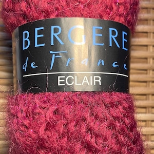 Peut inclure: Pelote de laine bordeaux avec une étiquette "BERGERE de France ECLAIR". La laine a une texture duveteuse avec des brins de paillettes. Idéale pour les projets de tricot et de crochet.