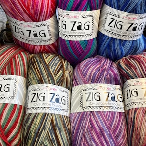 Könnte beinhalten: Sieben Knäuel King Cole Zig Zag 4ply Garn in verschiedenen bunten Farbverläufen. Jedes Knäuel hat ein weißes Papieretikett mit dem Text Zig Zag und 4ply suitable for socks. Das Garn zeigt mehrfarbige Muster in Rot-, Rosa-, Blau-, Lila-, Grün-, Braun- und Cremetönen mit einer gedrehten Faserstruktur.