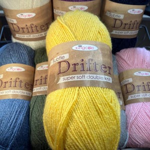 May include: A display of colorful yarn skeins, including yellow, blue, pink, and cream. Each skein is labeled with "Drifter" and "Super Soft Double Knit" in a brown label. The yarn is arranged in a wire basket.