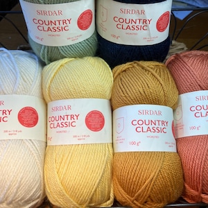 Könnte beinhalten: Eine Auswahl an Sirdar Country Classic-Wolle in Creme, Gelb, Gold, Orange, Grün und Marineblau. Jede 100g-Wollknäuel ist mit dem Markennamen und Details versehen.