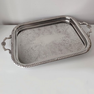Op de afbeelding: Een rechthoekig zilverkleurig dienblad met gedetailleerde gegraveerde krulpatronen in het midden, een opstaande rand met een kralenafwerking en twee decoratieve handgrepen met schelpachtige motieven aan weerszijden van het blad.