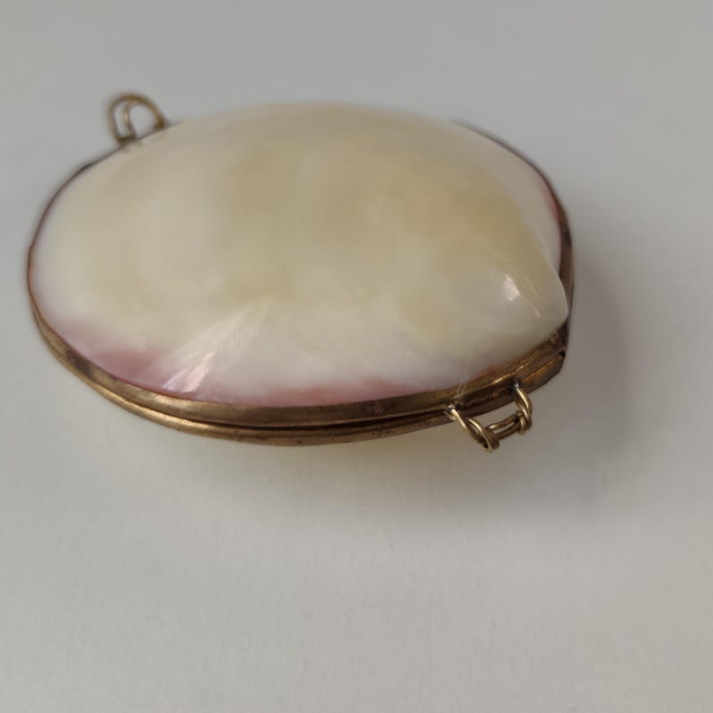 May include: An oval-shaped locket featuring a smooth, cream-colored shell inlay with subtle pinkish hues along the edges. The shell is encased in a metallic gold-toned frame with two small loops attached to the sides for connecting to a chain.