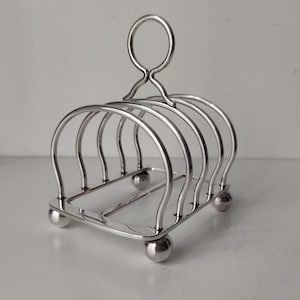 May include: A silver-toned metal toast rack featuring four arched slots for holding slices of bread. The structure includes a central circular handle at the top and is supported by four small, spherical ball feet at each corner of the rectangular base.