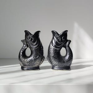 May include: A pair of matching black ceramic fish-shaped pitchers. Each vessel features a wide, open mouth at the top, a curved body with detailed scale textures, and a handle formed by the tail fin curving upward to meet the back of the head.