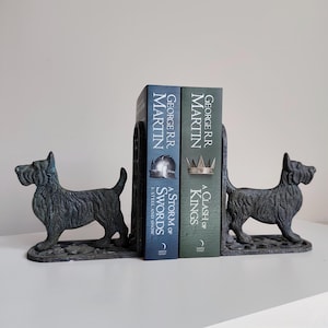 May include: A pair of cast iron bookends featuring a Scottish Terrier dog design. The dark gray bookends hold two books with the titles "A Storm of Swords" and "A Clash of Kings" by George R.R. Martin. The bookends are on a white surface.