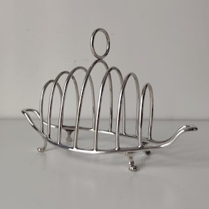 May include: A silver-toned metal toast rack featuring a series of arched wire dividers, a central circular handle at the top, and two small handles on either side. The structure is supported by four small, curved feet at the base.