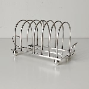 May include: A silver-toned metal toast rack featuring six arched wire slots for holding slices of bread. The structure is supported by four small spherical feet and includes two decorative curved handles on the ends, creating a functional kitchen accessory.