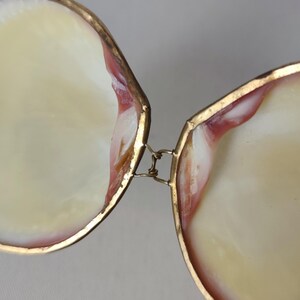 May include: A hinged box crafted from two iridescent seashell halves with smooth, cream-colored interiors and reddish-brown accents along the edges. The shells are joined by a thin, metallic gold-toned wire hinge that allows the box to open.