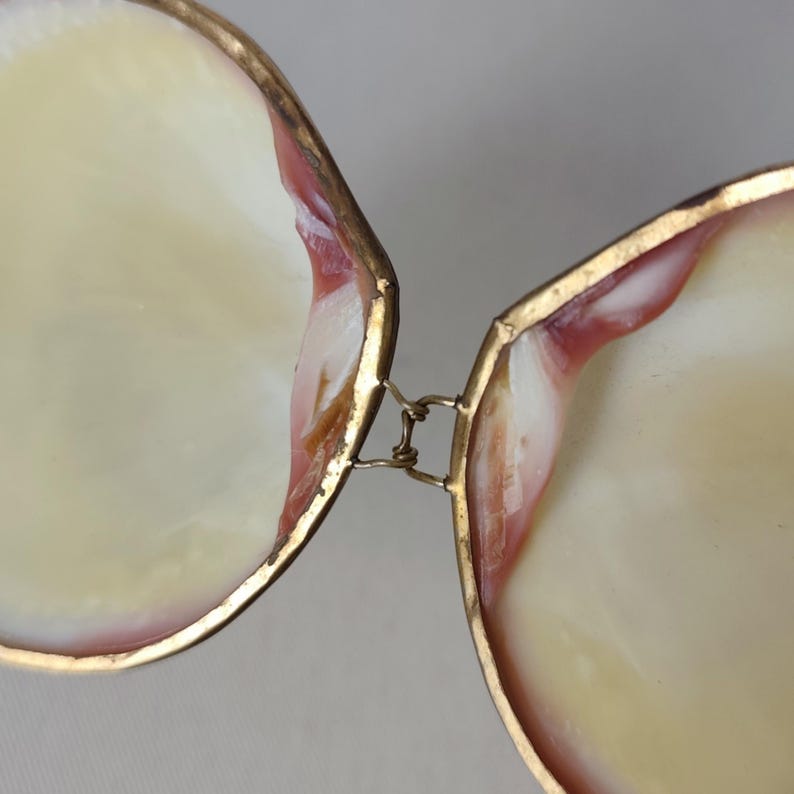 May include: A hinged box crafted from two iridescent seashell halves with smooth, cream-colored interiors and reddish-brown accents along the edges. The shells are joined by a thin, metallic gold-toned wire hinge that allows the box to open.