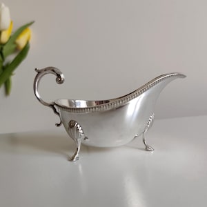 Puede incluir: Una salsera plateada con un asa curva y tres patas pequeñas. El recipiente tiene un borde decorativo y un pico. El diseño es elegante y clásico, adecuado para servir salsas o salsas.