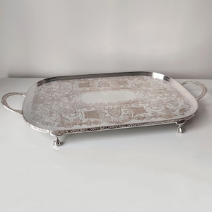 May include: A rectangular silver-toned serving tray with rounded corners, featuring intricate embossed floral and scrollwork patterns across the surface. The tray includes two curved side handles and is supported by four small, claw-style feet.