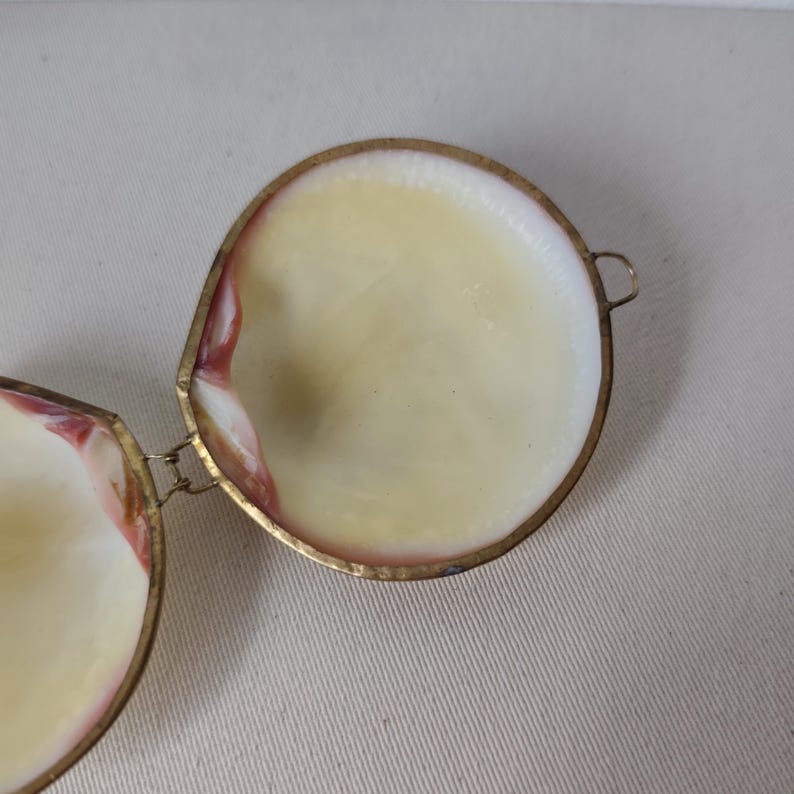 May include: A hinged circular container crafted from two natural seashells with a smooth, cream-colored interior and reddish-brown edges. The shells are joined by a thin, metallic gold-toned frame and a small hinge, featuring a small metal loop.