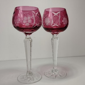 May include: Two wine glasses featuring deep magenta-coloured bowls with intricate cut-glass floral and geometric star patterns. Each glass has a clear, faceted stem and a circular base with a radiating starburst design etched into the glass.