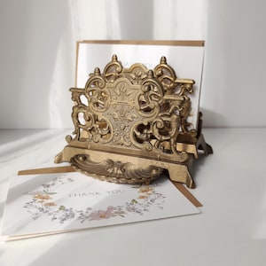 May include: A brass letter holder featuring an ornate, openwork metal design with scrollwork and floral motifs. The holder contains white envelopes, and a white card with a floral wreath illustration and the text THANK YOU rests in front.