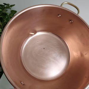 May include: A copper-coloured metal bowl with a rounded shape and a shiny finish. The bowl has a brass handle and rivets. The interior of the bowl is smooth and reflective, with a circular base. Likely used for cooking or serving.