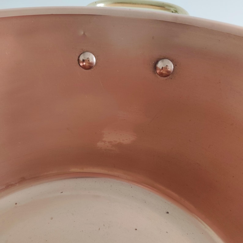 May include: Close-up of a copper cooking pot with a polished surface. Two silver rivets are visible on the side, and a gold-coloured handle is at the top. The pot has a traditional design, suitable for cooking.