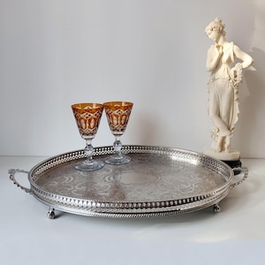 Puede incluir: Una bandeja de servicio ovalada grande de metal plateado con grabados ornamentales, un borde calado y dos asas decorativas, acompañada de dos copas de vino de cristal ámbar con un patrón de diamantes tallados en el cáliz.