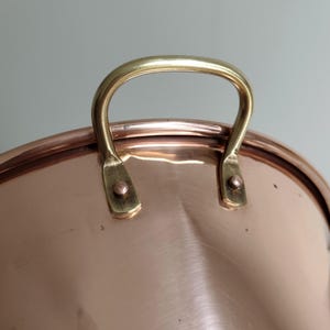 May include: Close-up of a copper pot with a brass handle. The pot has a shiny, reflective surface and a curved rim. The brass handle is arched and attached to the pot with rivets. The pot is likely used for cooking.