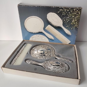 Può includere: Un set da toeletta in scatola contenente uno specchio a mano, una spazzola per capelli e un pettine color argento. Lo specchio e la spazzola presentano intricati motivi floreali in rilievo sui manici e sul retro. Il pettine ha un corpo color crema.