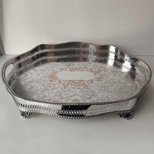 May include: A silver-plated serving tray with an oval shape, featuring an intricate engraved floral pattern on the base, a pierced gallery rim, two integrated side handles, and four decorative feet. The center has a smooth, blank area.