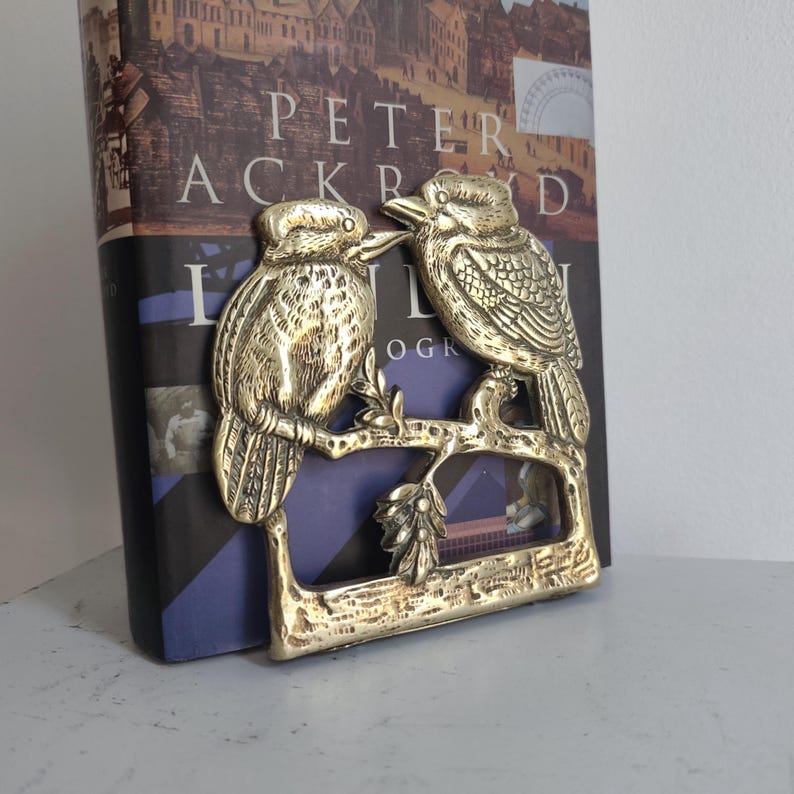 May include: A polished brass decorative object featuring two kookaburras perched on a textured branch with leaves. The metal sculpture has detailed engravings of feathers and facial features, with a rectangular base and a sturdy frame structure.