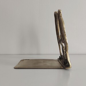 May include: A brass bookend featuring a vertical, openwork decorative panel attached to a flat, horizontal base. The metal has a polished, reflective gold-toned finish with visible screws connecting the upright section to the base plate.