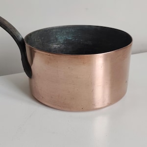 May include: A copper saucepan with a dark handle. The pot has a rounded shape and a shiny, reflective surface. The interior of the pot is a darker colour, contrasting with the copper exterior. The handle is attached to the side of the pot.