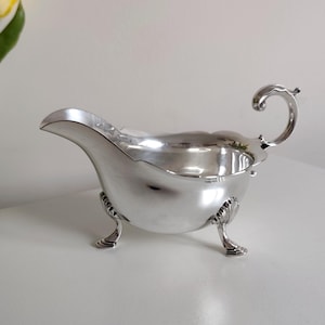 Puede incluir: Una salsera plateada con un pico curvo y un asa decorativa. La salsera tiene un cuerpo redondeado y se apoya en tres patas ornamentadas. La superficie pulida refleja la luz, resaltando su elegante diseño. Una pieza de servicio clásica.