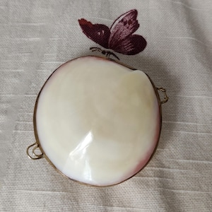 May include: A round, smooth, cream-colored shell box with a polished finish and a metallic gold-toned rim. The container features small metal loops on the sides and is positioned against a white fabric background with a dark purple butterfly embroidery.