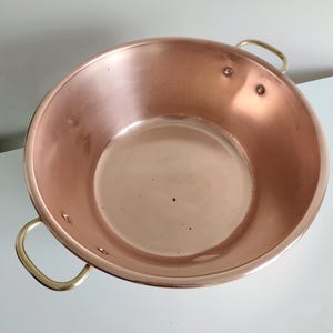 May include: A copper pan with brass handles. The pan is round with a wide, shallow bowl and two curved brass handles attached to the sides. The copper has a shiny, reflective surface.