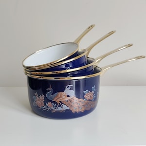 May include: Set of four navy blue enamel saucepans with gold-colored handles and rims. Each pan features a peacock and floral design in shades of brown, peach, and blue. The pans are stacked, showcasing their varying sizes and shapes.