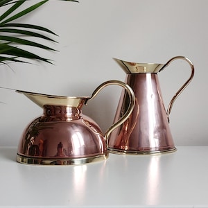 May include: Two copper and brass pitchers, one with a rounded body and the other with a conical body. Both pitchers have a shiny, polished finish.